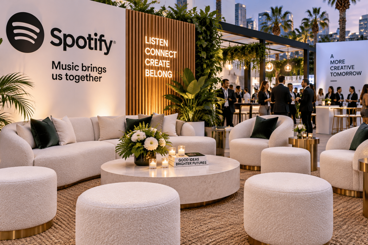 Branded event lounge with rental sofas, chairs, ottomans, cocktail tables, greenery, and guests networking in a premium event environment.
